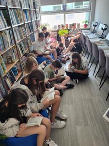 Na zdjęciu znajduje się młodzież czytająca książki. Młodzież siedzi - w tle księgozbiór biblioteki.