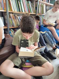 Na zdjęciu znajduje się młodzież czytająca książki. Młodzież siedzi - w tle księgozbiór biblioteki.