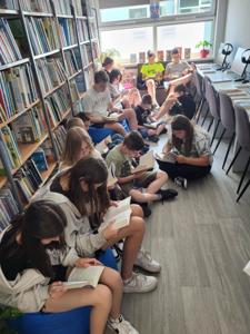 Na zdjęciu znajduje się młodzież czytająca książki. Młodzież siedzi - w tle księgozbiór biblioteki.