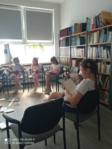 Na zdjęciu znajduje się młodzież czytająca książki. Młodzież siedzi - w tle księgozbiór biblioteki.