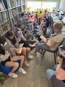 Na zdjęciu znajduje się młodzież czytająca książki. Młodzież siedzi - w tle księgozbiór biblioteki.