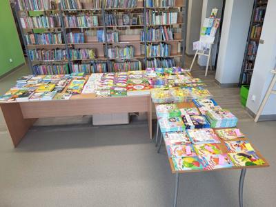 Na zdjęciu znajdują się książki poukładane na stołach. W tle księgozbiór biblioteki. Zdjęcie wykonano w bibliotece.