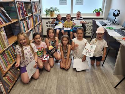 Na zdjęciu znajdują się uczestnicy warsztatów. Młodzież trzyma w rękach prace plastyczne. Zdjęcie wykonano w bibliotece.