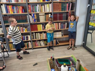 Na zdjęciu znajdują się uczestnicy zajęć. Uczestnicy wykonują zadania. W tle księgozbiór biblioteki.  Zdjęcie wykonano w bibliotece.