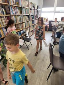 Na zdjęciu znajdują się uczestnicy zajęć. Dzieci wykonują powierzone im zadania. W tle księgozbiór biblioteki.