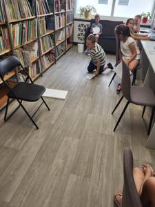 Na zdjęciu znajdują się uczestnicy zajęć. Dzieci wykonują powierzone im zadania. W tle księgozbiór biblioteki.
