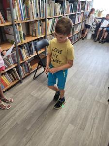 Na zdjęciu znajdują się uczestnicy zajęć. Dzieci wykonują powierzone im zadania. W tle księgozbiór biblioteki.