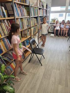 Na zdjęciu znajdują się uczestnicy zajęć. Dzieci wykonują powierzone im zadania. W tle księgozbiór biblioteki.