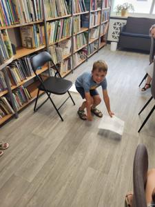 Na zdjęciu znajdują się uczestnicy zajęć. Dzieci wykonują powierzone im zadania. W tle księgozbiór biblioteki.
