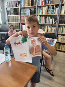 Na zdjęciu się uczestnik zajęć. Chłopiec trzyma rękach prace plastyczne. Zdjęcie wykonano w bibliotece.