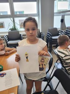 Na zdjęciu się uczestniczka zajęć. Dziewczynka trzyma rękach prace plastyczne. Zdjęcie wykonano w bibliotece.