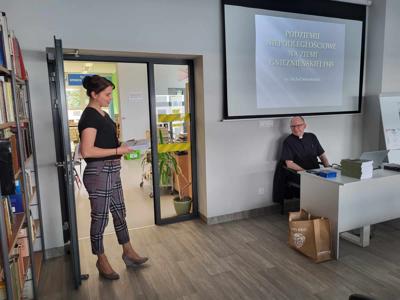 Na zdjęciu znajdują się prelegenci. W tle wyświetlana prezentacja multimedialna. Zdjęcie wykonano w bibliotece.