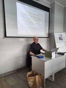 Na zdjęciu znajduje się prelegent. W tle wyświetlana prezentacja multimedialna. Zdjęcie wykonano w bibliotece.