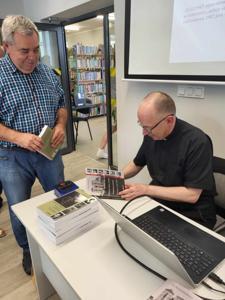 Na zdjęciu znajduje się prelegent siedzący przy biurku i uczestnicy spotkania. Prelegent podpisuje książkę. Zdjęcie wykonano w bibliotece.