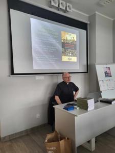 Na zdjęciu znajduje się prelegent. W tle wyświetlana prezentacja multimedialna. Zdjęcie wykonano w bibliotece.