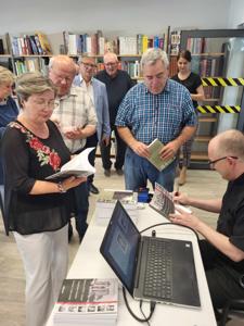 Na zdjęciu znajduje się prelegent siedzący przy biurku i uczestnicy spotkania. Prelegent podpisuje książkę. Zdjęcie wykonano w bibliotece.