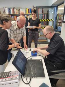 Na zdjęciu znajduje się prelegent siedzący przy biurku i uczestnicy spotkania. Prelegent podpisuje książkę. Zdjęcie wykonano w bibliotece.