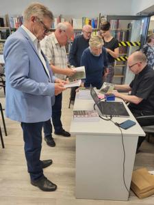 Na zdjęciu znajduje się prelegent siedzący przy biurku i uczestnicy spotkania. Prelegent podpisuje książkę. Zdjęcie wykonano w bibliotece.