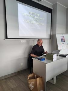 Na zdjęciu znajduje się prelegent. W tle wyświetlana prezentacja multimedialna. Zdjęcie wykonano w bibliotece.