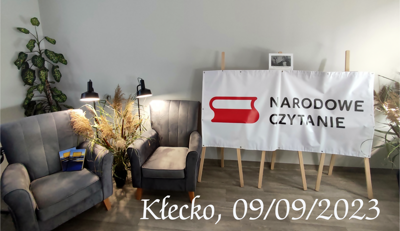 Na zdjęciu znajduje się wystawa Narodowego Czytania - książki, fotele, bilbord, lampy oraz kwiaty.