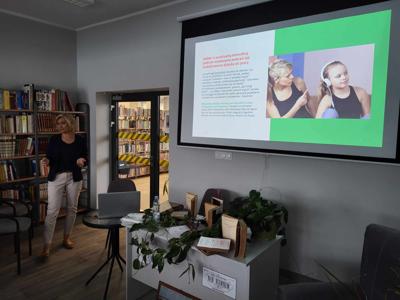 Na zdjęciu znajduje się osoba prowadząca wykład. W tle księgozbiór biblioteki oraz prezentacja multimedialna. Zdjęcie wykonano w bibliotece.