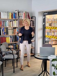 Na zdjęciu znajduje się osoba prowadząca wykład. W tle księgozbiór biblioteki. Zdjęcie wykonano w bibliotece.
