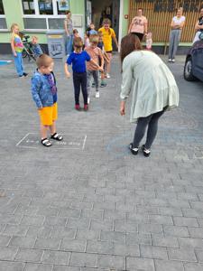 Na zdjęciu znajdują się uczestnicy warsztatów. Dzieci stoją i wykonują zadania. Zdjęcie wykonano na parkingu przy bibliotece.