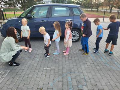 Na zdjęciu znajdują się uczestnicy warsztatów. Dzieci stoją i wykonują zadania. Zdjęcie wykonano na parkingu przy bibliotece.