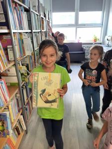 Na zdjęciu znajdują się uczestnicy zajęć. Dziewczynka trzyma w ręce kartkę. Zdjęcie wykonano w bibliotece.