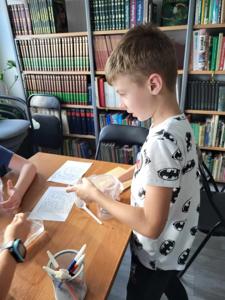 Na zdjęciu znajdują się uczestnicy zajęć. Dzieci wykonują zadanie przy stołach. Zdjęcie wykonano w bibliotece.