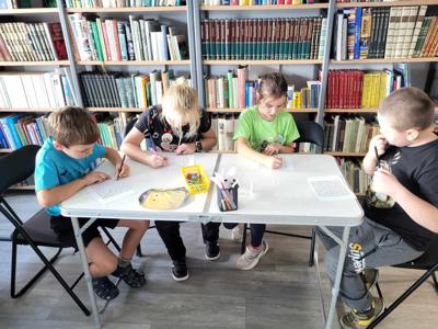 Na zdjęciu znajdują się uczestnicy zajęć. Dzieci wykonują zadanie przy stołach. Zdjęcie wykonano w bibliotece.