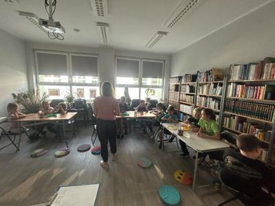 Na zdjęciu znajdują się uczestnicy zajęć. Dzieci wykonują zadanie przy stołach. Zdjęcie wykonano w bibliotece.