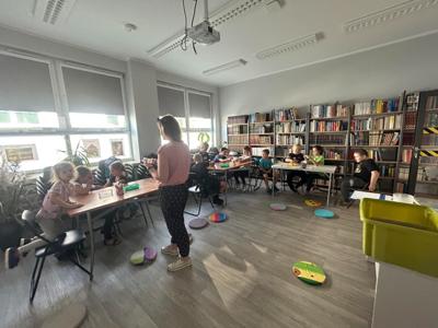 Na zdjęciu znajdują się uczestnicy zajęć. Dzieci wykonują zadanie przy stołach. Zdjęcie wykonano w bibliotece.
