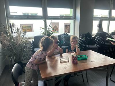 Na zdjęciu znajdują się uczestnicy zajęć. Dzieci wykonują zadanie przy stołach. Zdjęcie wykonano w bibliotece.