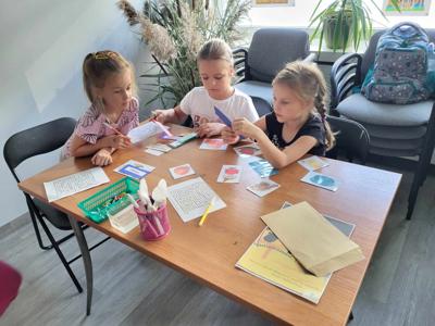 Na zdjęciu znajdują się uczestnicy zajęć. Dzieci wykonują zadanie przy stołach. Zdjęcie wykonano w bibliotece.