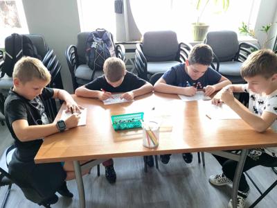 Na zdjęciu znajdują się uczestnicy zajęć. Dzieci wykonują zadanie przy stołach. Zdjęcie wykonano w bibliotece.