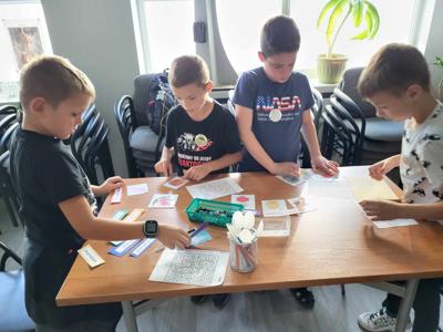 Na zdjęciu znajdują się uczestnicy zajęć. Dzieci wykonują zadanie przy stołach. Zdjęcie wykonano w bibliotece.