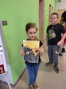 Na zdjęciu znajdują się uczestnicy zajęć. Dziewczynka trzyma w ręce kartkę. Zdjęcie wykonano w bibliotece.