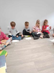 Na zdjęciu znajdują się uczestnicy zajęć. Dzieci wykonują zadania siedząc na poduszkach. Zdjęcie wykonano w bibliotece.