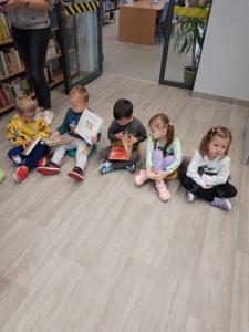 Na zdjęciu znajdują się uczestnicy zajęć. Dzieci wykonują zadania siedząc na poduszkach. Zdjęcie wykonano w bibliotece.