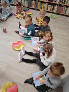 Na zdjęciu znajdują się uczestnicy zajęć. Dzieci wykonują zadania siedząc na poduszkach. Zdjęcie wykonano w bibliotece.