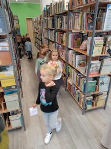 Na zdjęciu znajdują się uczestnicy zajęć. Dzieci wykonują zadania pomiędzy regałami z książkami.  Zdjęcie wykonano w bibliotece.