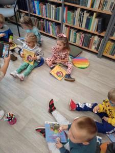 Na zdjęciu znajdują się uczestnicy zajęć. Dzieci wykonują zadania siedząc na poduszkach. Zdjęcie wykonano w bibliotece.