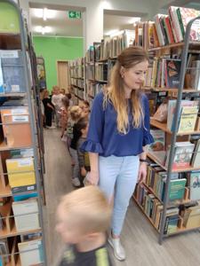 Na zdjęciu znajdują się uczestnicy zajęć. Dzieci wykonują zadania pomiędzy regałami z książkami.  Zdjęcie wykonano w bibliotece.