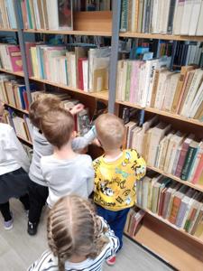 Na zdjęciu znajdują się uczestnicy zajęć. Dzieci wykonują zadania pomiędzy regałami z książkami.  Zdjęcie wykonano w bibliotece.