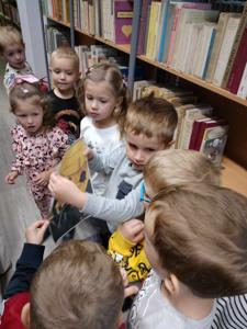 Na zdjęciu znajdują się uczestnicy zajęć. Dzieci wykonują zadania pomiędzy regałami z książkami.  Zdjęcie wykonano w bibliotece.