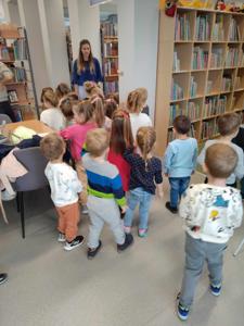 Na zdjęciu znajdują się uczestnicy zajęć. Dzieci wykonują zadania pomiędzy regałami z książkami.  Zdjęcie wykonano w bibliotece.