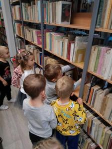 Na zdjęciu znajdują się uczestnicy zajęć. Dzieci wykonują zadania pomiędzy regałami z książkami.  Zdjęcie wykonano w bibliotece.
