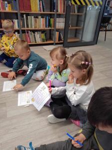 Na zdjęciu znajdują się uczestnicy zajęć. Dzieci wykonują zadania siedząc na poduszkach. Zdjęcie wykonano w bibliotece.