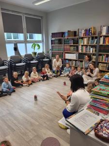 Na zdjęciu znajdują się uczestnicy zajęć. Dzieci wykonują zadania siedząc na poduszkach. Zdjęcie wykonano w bibliotece.